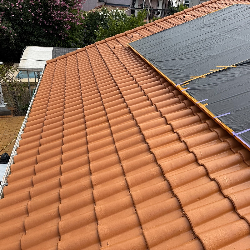 Toiture en tuiles de terre cuite orange. Une large zone est temporairement couverte par une bâche noire pour travaux de rénovation ou protection.